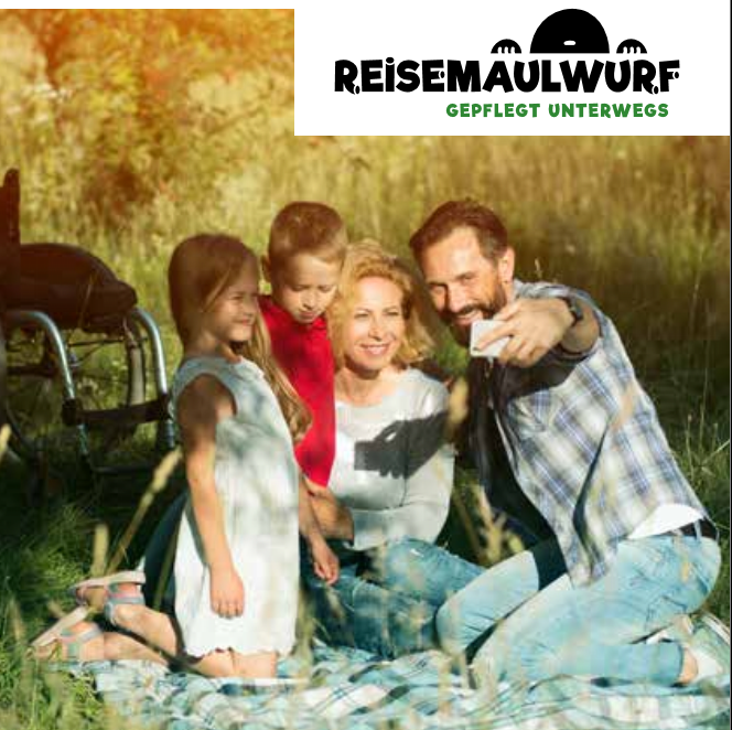 Eine Familie beim Picknick im Sonnenschein auf einer Decke. Im Hintergrund ist ein Rollstuhl zu sehen